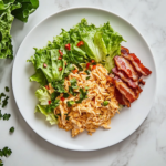 A white ceramic plate on a white marble cooktop is beautifully plated with creamy curry chicken salad, crisp lettuce wraps, and crispy bacon slices on the side, ready to be served.