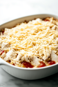 A baking dish filled with the turkey mixture, evenly spread out. Freshly shredded Gruyère cheese is being sprinkled generously over the top, creating a thick, cheesy layer before it goes into the oven.