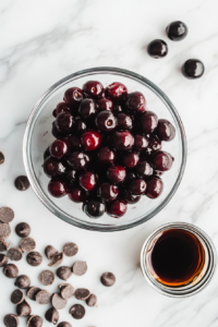 Close-up shot of a bowl of drained black cherries and fresh cherries on the white marble cooktop, with chocolate chips and a small cup of kirsch liquid beside them. The cherries glisten slightly