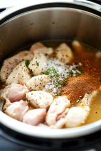 A close-up of boneless, skinless chicken being placed into the Instant Pot with rich, golden chicken broth. Onion powder, garlic powder, sea salt, and fresh cracked black pepper are sprinkled on top, adding layers of savory flavor before cooking.