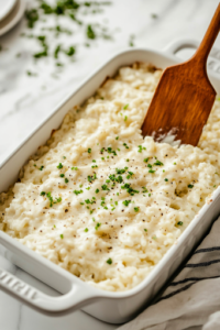 A greased 9×13-inch glass baking dish sits on the white marble cooktop, with the creamy chicken and rice mixture being spread evenly with a spatula. The surface is smooth and level, ready for the next layer.