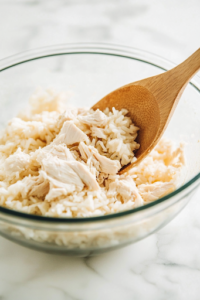 The same glass mixing bowl now contains diced cooked chicken and cooked rice, added to the creamy sauce. A wooden spoon is gently folding the ingredients together, ensuring an even, thick, and well-mixed filling.