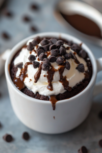 A chocolate mug cake topped with melted chocolate chips, a swirl of whipped cream, and a drizzle of chocolate sauce. A spoon rests beside the mug, ready to dig into the decadent dessert.