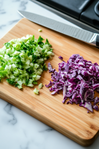 A close-up shot of a wooden cutting board on a white marble cooktop, showcasing finely chopped green and red cabbage neatly arranged in separate piles. A sharp chef’s knife is positioned beside the cabbage, prepared for additional chopping.