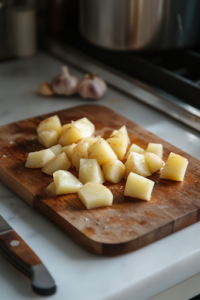 A close-up shot of a wooden cutting board on the white marble cooktop, with peeled russet potatoes chopped into equal-sized chunks. A chef’s knife rests beside a few whole garlic cloves, all prepped and ready to be added to the slow cooker.