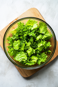 An overhead shot of a wooden cutting board with freshly chopped, vibrant green romaine lettuce, placed in a glass salad bowl. The bright lettuce contrasts beautifully against the neutral wooden background.