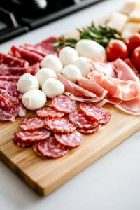 A close-up of a wooden cutting board on a white marble cooktop, with a sharp knife slicing salami, prosciutto, and fresh mozzarella balls into bite-sized pieces, neatly arranged and ready for the salad.