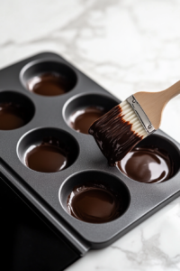 A patty pan on the white marble cooktop, with two teaspoons of melted chocolate spooned in. A small pastry brush is gently spreading the chocolate up the sides and bottom of the pan.