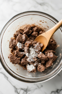 A close-up shot of a glass mixing bowl resting on the white marble cooktop, filled with chunks of beef stew meat coated in all-purpose flour, salt, and black pepper. A wooden spoon gently stirs, ensuring each piece is evenly covered in the seasoned flour for a perfect sear.