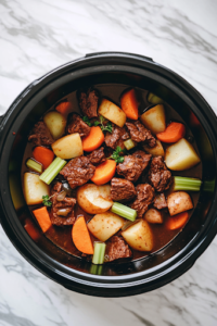 A close-up shot of a black slow cooker sitting on the white marble cooktop, filled with freshly diced potatoes, sliced carrots, chopped celery, and onions. The vibrant colors of the vegetables contrast beautifully against the floured beef, creating a hearty base for the stew.