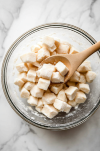 A glass mixing bowl on a white marble cooktop filled with cubed chicken coated in cornstarch, salt, and pepper. A wooden spoon gently stirs the ingredients, ensuring an even coating on the chicken pieces before cooking.