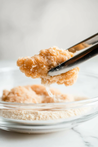 : A coated piece of chicken being lifted from the buttermilk with tongs over a glass bowl on a white marble cooktop. The excess buttermilk drips off before the chicken is placed into the seasoned flour mixture for coating.