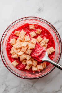A mixing bowl with red frosting and white chocolate-coated Chex cereal being stirred with a spatula.