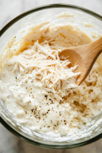 The same glass mixing bowl on the white marble cooktop, now containing shredded chicken and cooked rice. A wooden spoon is folding the ingredients together, ensuring everything is well combined into a rich and hearty mixture.
