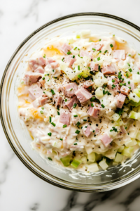 A close-up shot of the same glass mixing bowl on the white marble cooktop, now filled with the finely chopped ham, egg, onion, and celery added to the creamy dressing. A wooden spoon is partially submerged, preparing to mix everything together.