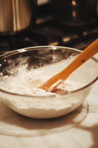 A close-up shot of the glass mixing bowl on the white marble cooktop as the dry ingredients are gradually incorporated into the wet mixture. A spatula is gently folding the dough together, ensuring even distribution without overmixing.
