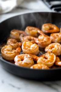 A close-up shot of the cooked shrimp resting in the black skillet over a white marble cooktop. The shrimp have a deep golden hue, with steam rising and a wooden spatula ready for serving, indicating they are perfectly cooked and infused with flavors.