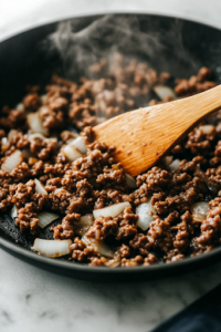 A close-up shot of a black skillet on the white marble cooktop, sizzling with lean ground beef, chopped onion, and minced garlic. The meat browns evenly as steam rises, while a wooden spatula stirs the mixture, releasing a rich, savory aroma.
