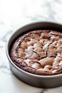 A close-up shot of the freshly baked chocolate cake in the pan on the white marble cooktop, cooling for 10 minutes. The cake looks moist and slightly domed, indicating a soft and fluffy texture.