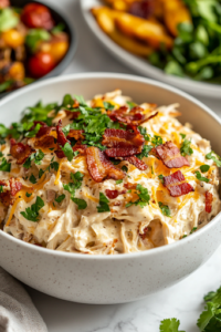 A close-up of a white serving bowl filled with creamy, cheesy shredded chicken, generously topped with crispy bacon and fresh herbs. A side of roasted vegetables and a fresh green salad sit beside it on a rustic wooden table, making for a comforting and satisfying meal.