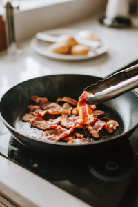 A close-up of a black skillet on a white marble cooktop, sizzling with crispy bacon strips that are turning golden brown. A pair of tongs lifts one piece while a nearby paper towel-lined plate is ready for draining excess grease.