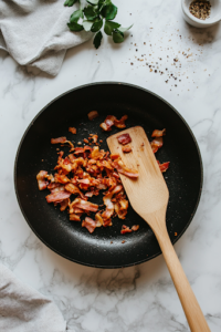 A close-up of a black skillet on a white marble cooktop, sizzling with crispy bacon strips turning golden brown. A wooden spatula rests on the side, ready to crumble the bacon into small pieces.