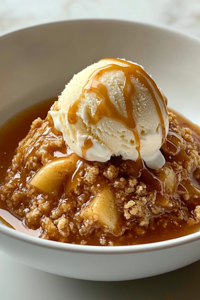 A bowl with a warm serving of slow-cooked apple crisp on a wooden countertop. A scoop of melting vanilla ice cream is on top, drizzled with caramel sauce. A spoon rests beside the bowl.