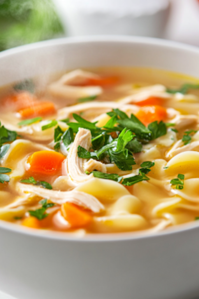 A close-up shot of a steaming bowl of homemade chicken noodle soup placed on a rustic wooden table. The soup is garnished with fresh parsley, enhancing its vibrant color. It is served alongside a plate of Brussels sprouts and salted caramel chocolate chip cookies, creating a cozy and comforting meal.