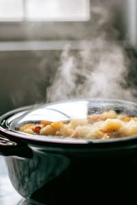A close-up shot of a crockpot covered with a glass lid as the cheesy hashbrown mixture cooks. Steam slightly fogs up the inside of the lid, indicating the cooking process. The white marble cooktop beneath reflects the kitchen’s bright and neat ambiance.