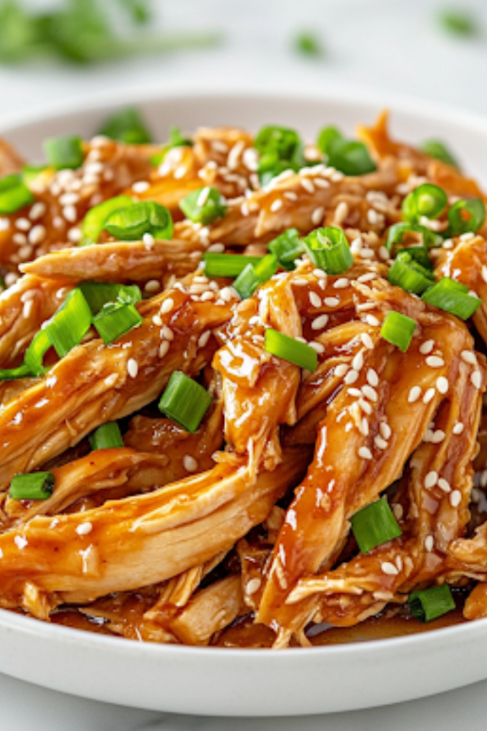 A white plate on a white marble cooktop, filled with shredded honey garlic chicken served over cauliflower rice. The dish is garnished with toasted sesame seeds and freshly chopped green onions, adding a final touch of flavor and color, ready to be enjoyed.