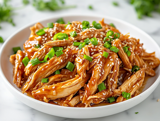 A white plate on a white marble cooktop, filled with shredded honey garlic chicken served over cauliflower rice. The dish is garnished with toasted sesame seeds and freshly chopped green onions, adding a final touch of flavor and color, ready to be enjoyed.