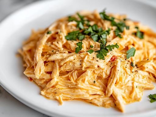 A white plate on a white marble cooktop, featuring a generous serving of creamy shredded Italian chicken over pasta, garnished with fresh herbs, ready to be enjoyed.