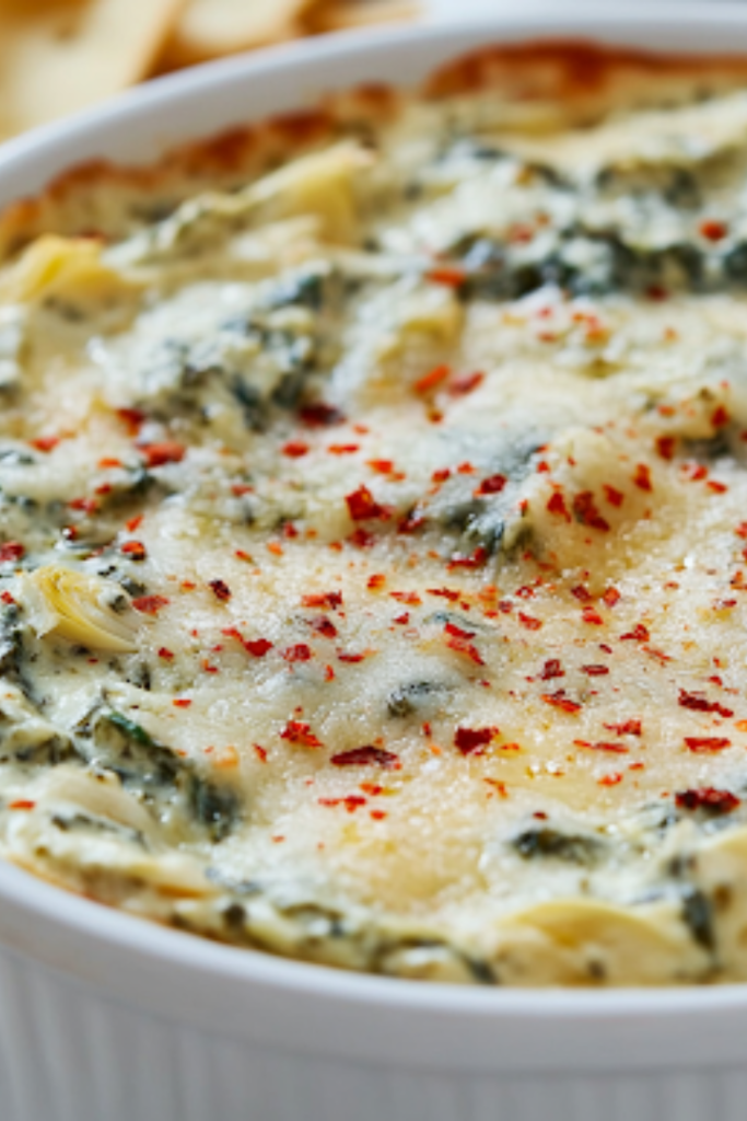 A serving dish filled with creamy spinach artichoke dip, garnished with extra Parmesan and red pepper flakes. The dish is surrounded by crispy toasted baguette slices and fresh veggie sticks, beautifully arranged on the white marble countertop, making it a perfect appetizer for sharing.