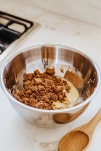 A glass mixing bowl filled with crushed Biscoff biscuits and melted butter. A spoon is seen stirring the mixture before pressing it into a round tin.