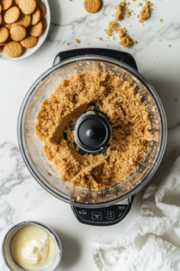 A food processor filled with crushed graham crackers being pulsed into fine crumbs. A small bowl with melted butter is ready to be mixed in.