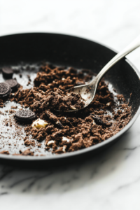 Close-up shot of a crushed Oreos, instant espresso powder, and melted butter. The mixture is being pressed into the bottom of a loaf pan, creating a smooth crust base.