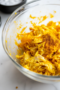 Curry powder is being sprinkled over the shredded chicken and mayo mixture in a glass mixing bowl on the white marble cooktop. A small dish of salt and pepper sits nearby, ready for seasoning.