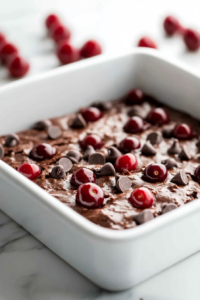 Close-up shot of freshly baked brownies cooling in the tray on the white marble cooktop. Melted dark chocolate is being drizzled over the top, and delicate chocolate curls are sprinkled for decoration
