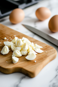 A close-up shot of a wooden cutting board on a white marble cooktop, with hard-boiled eggs neatly diced. A sharp knife rests beside the diced eggs, with eggshell fragments scattered in the background.