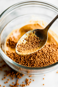 A close-up shot of a spoon adding ¼ teaspoon of creamy Dijon mustard into the glass mixing bowl on the white marble cooktop, bringing a tangy and slightly spicy depth to the Greek salad dressing.