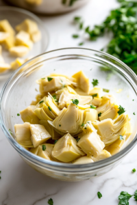 A close-up of a can of artichoke hearts being drained over a glass bowl on a white marble cooktop, with freshly chopped artichoke pieces set aside, ready to be mixed into the salad.