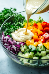 A side-angle shot of a large mixing bowl as a creamy dressing is poured over the vibrant salad ingredients. A hand holding a wooden spoon is about to toss everything together for even coating.