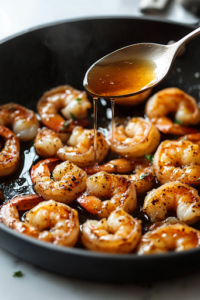 A close-up shot of sugar-free honey drizzling from a spoon over the seasoned shrimp in a black skillet over a white marble cooktop. The shrimp glisten as they absorb the rich, golden sweetness, enhancing their flavor and texture.