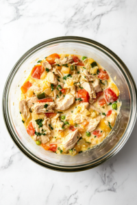 A smooth and creamy whisked mixture of eggs and heavy cream in a glass bowl, with visible flecks of seasoning. A hand is pouring it gently into the turkey and vegetable mixture, ensuring a rich and cohesive texture.