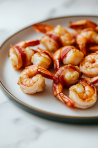 Close-up shot of a plate on the white marble cooktop, holding perfectly golden bacon-wrapped shrimp. A crispy texture is visible on the bacon, and the shrimp tails peek out invitingly.