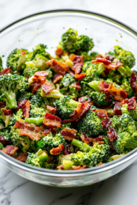 A glass mixing bowl on a white marble cooktop, filled with freshly tossed broccoli bacon salad, covered and ready for refrigeration. The dressing evenly coats the ingredients, with vibrant colors peeking through for a delicious final look.