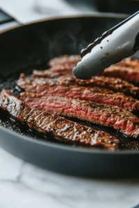 A close-up of a black skillet on a white marble cooktop, where a seasoned flank steak sizzles, forming a deep caramelized crust. A pair of tongs is flipping the steak, revealing a beautifully seared surface.
