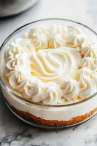 Close-up shot of whipped cream being gently folded into a smooth cream cheese mixture in a clear glass bowl on the white marble cooktop. A silicone spatula moves in delicate swirls, creating a light, airy texture.