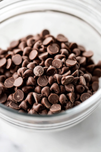 A close-up shot of chocolate chips being gently folded into the cookie dough inside the glass mixing bowl on the white marble cooktop. The spatula swirls through the dough, ensuring the chocolate chips are evenly distributed in every bite.