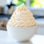A bowl filled with perfectly whipped French buttercream sits on a white marble cooktop. A spatula lifts a smooth, silky swirl, showing its luscious texture, ready for frosting.